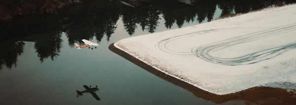 A small red-and-white plane reflects on the still surface of a frozen Alaskan lake, surrounded by snow and evergreen trees. This image captures the lifeline aviation provides in Alaska's vast, roadless wilderness.