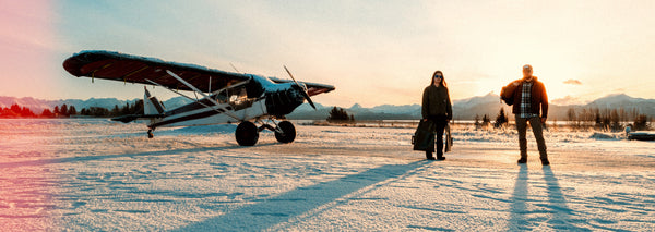 Two Alaskan pilots stand on a snow-covered runway next to a vintage plane, representing Randolph sunglasses' timeless craftsmanship and resilience for winter exploration.